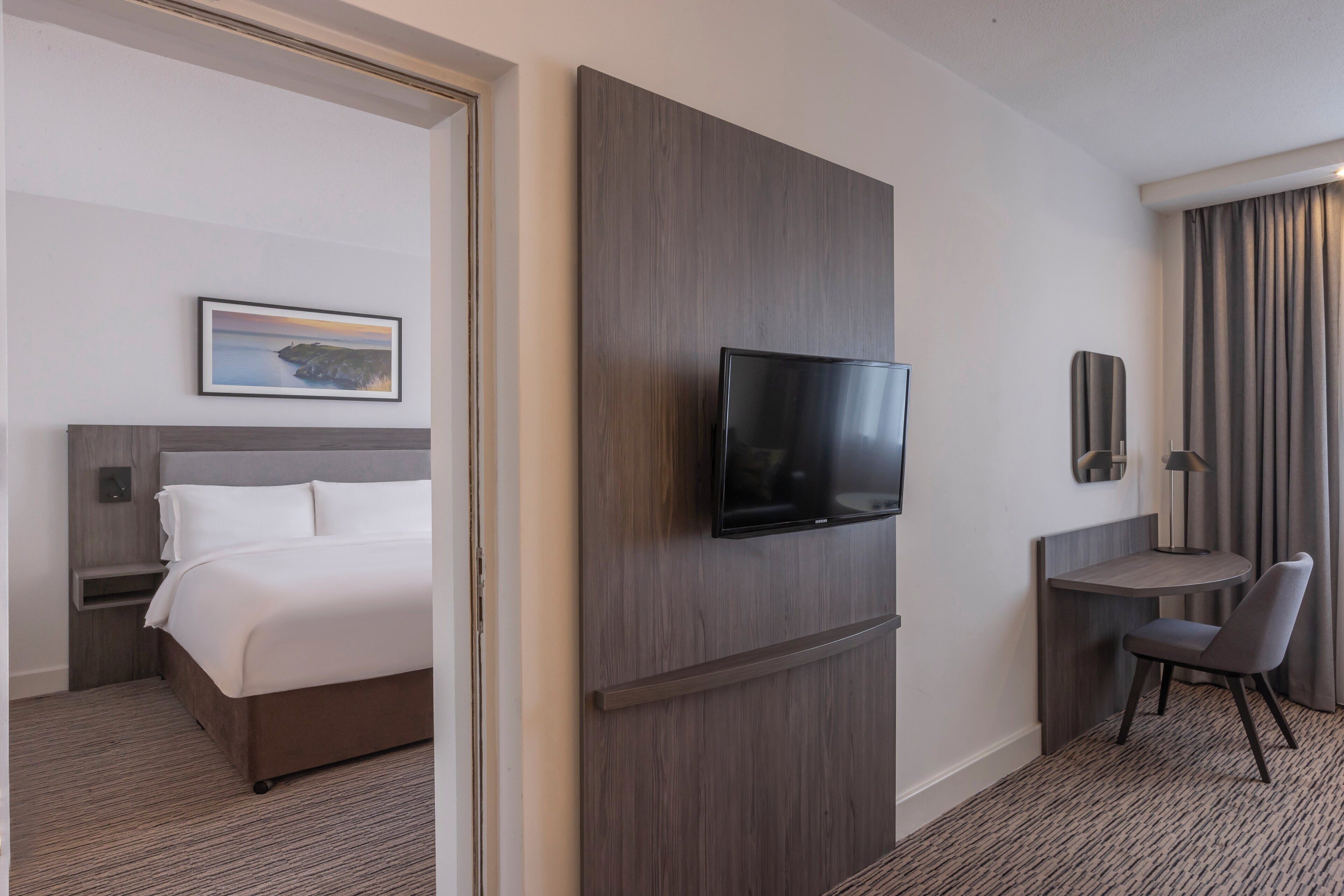 Una habitación de hotel con una cama, televisión instalada en la pared, un escritorio con silla y un ventanal con cortinas.