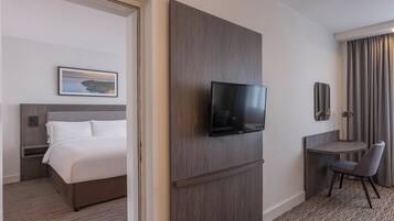 Una habitación de hotel con una cama, televisión instalada en la pared, un escritorio con silla y un ventanal con cortinas.
