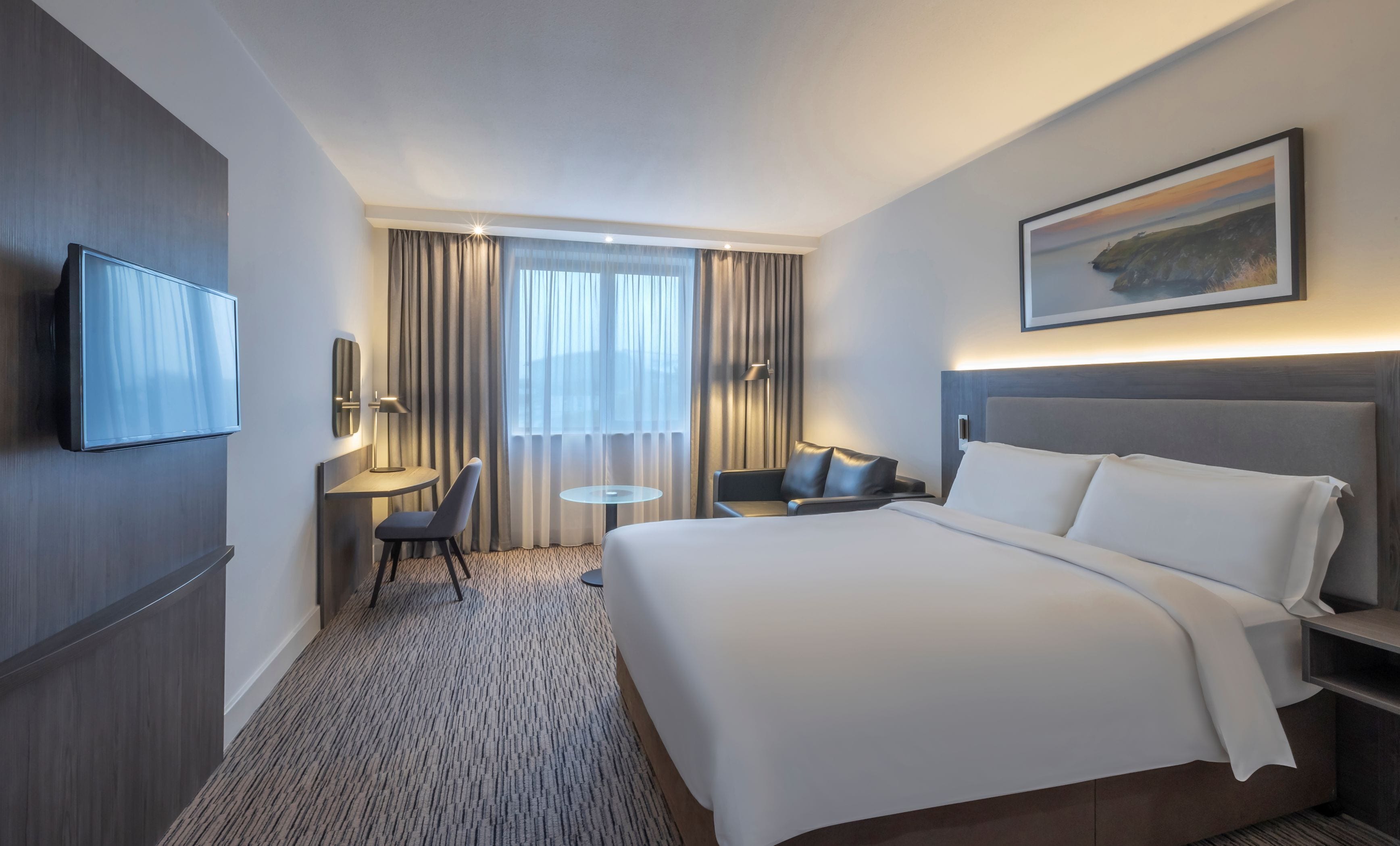 Ein Hotelzimmer mit einem großen Bett, einem flachbildfernseher an der Wand, einem Schreibtisch mit Stuhl, einem kleinen runden Tisch und einem Gemälde an der Wand.