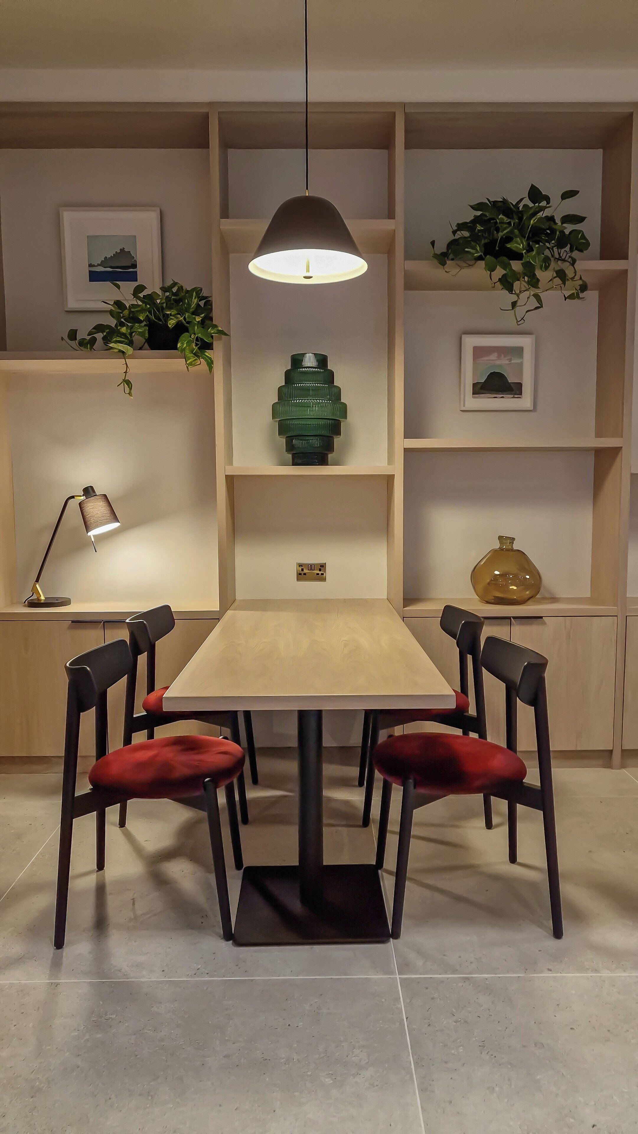 Un comedor con mesa de madera, cuatro sillas con cojines rojos, una lámpara colgante y estantes con plantas y adornos.