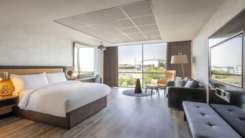 A modern hotel room with a large bed, a seating area with a sofa and armchairs, a small table, and a view of the cityscape through large windows.