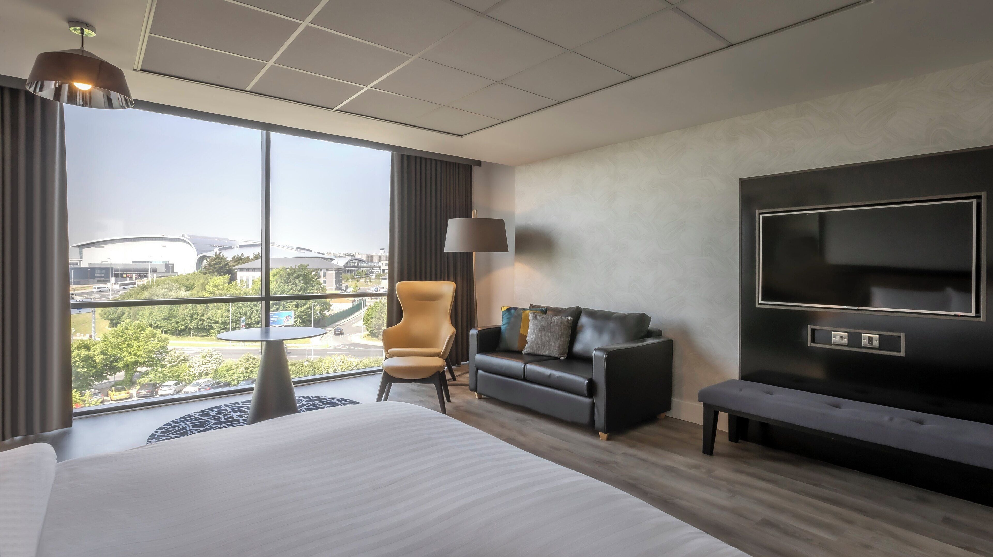 A hotel room with a large bed, a sofa, a chair, a small table, a TV, and a view of the city through large windows.