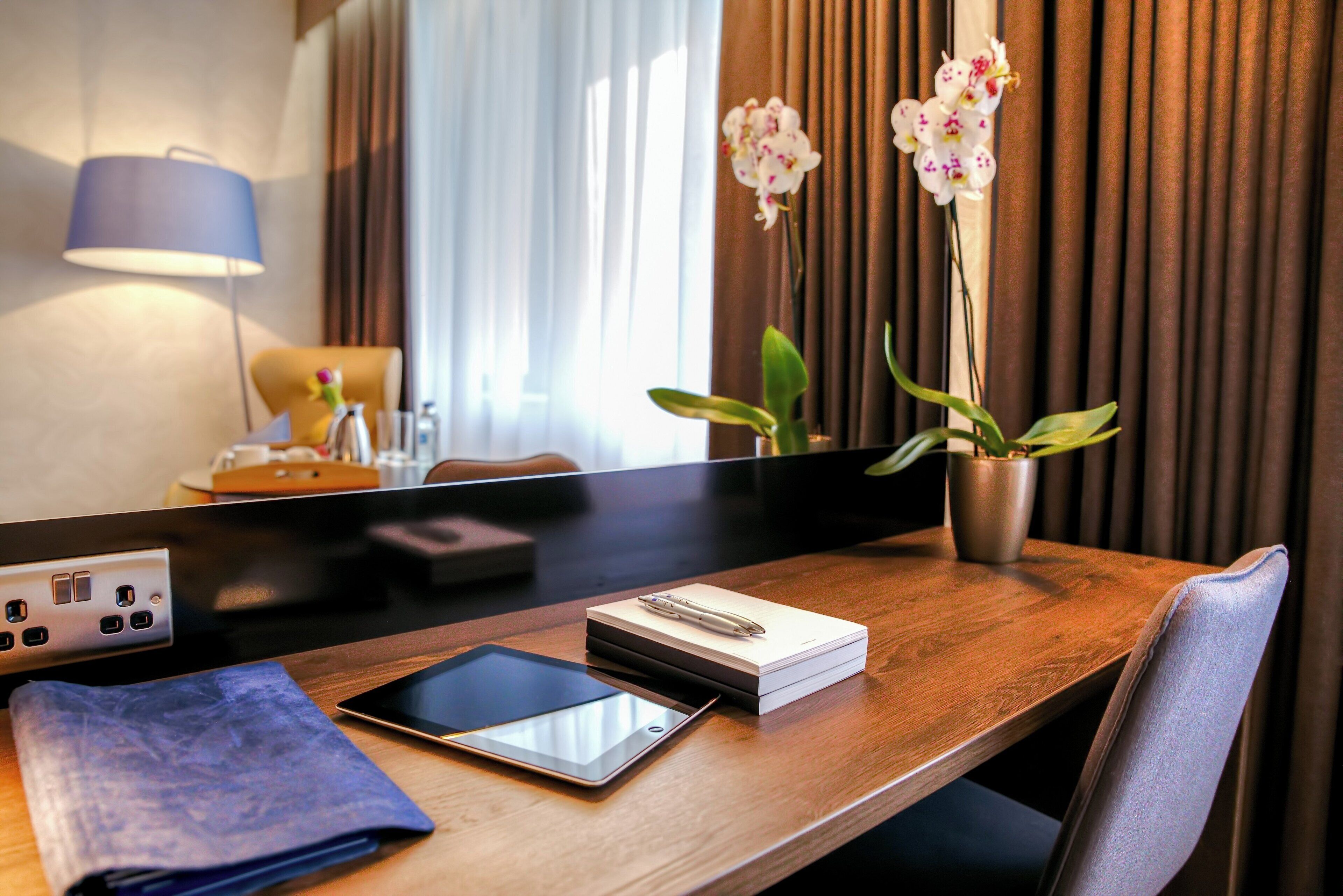 Habitación de hotel con un escritorio de madera, una tableta, un cuaderno, un bolígrafo y una orquídea en maceta.