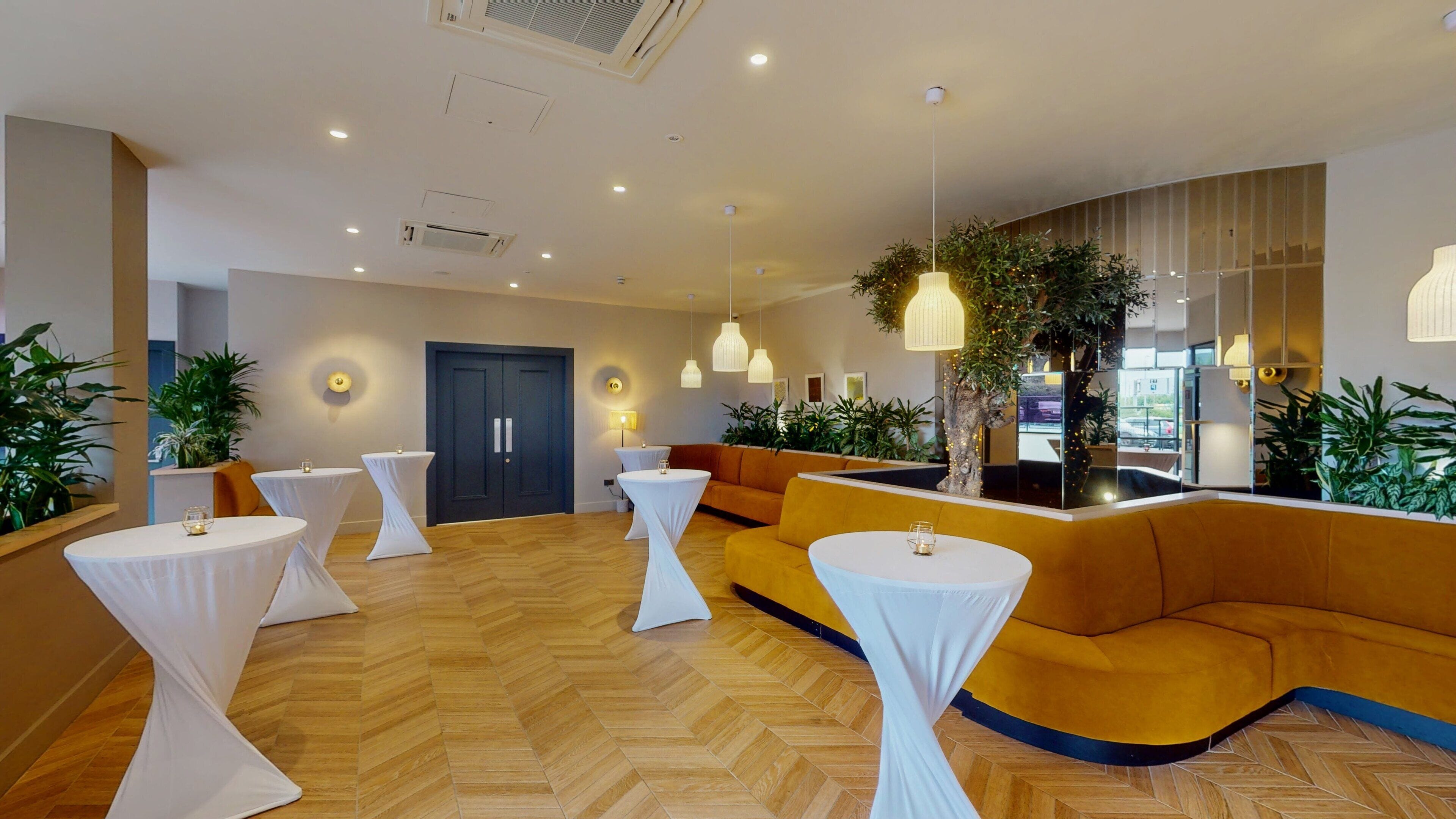 A modern hotel lobby with a herringbone wood floor, a central seating area with a yellow sofa, and multiple white tables with draped tablecloths.
