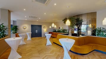 A modern hotel lobby with a herringbone wood floor, a central seating area with a yellow sofa, and multiple white tables with draped tablecloths.