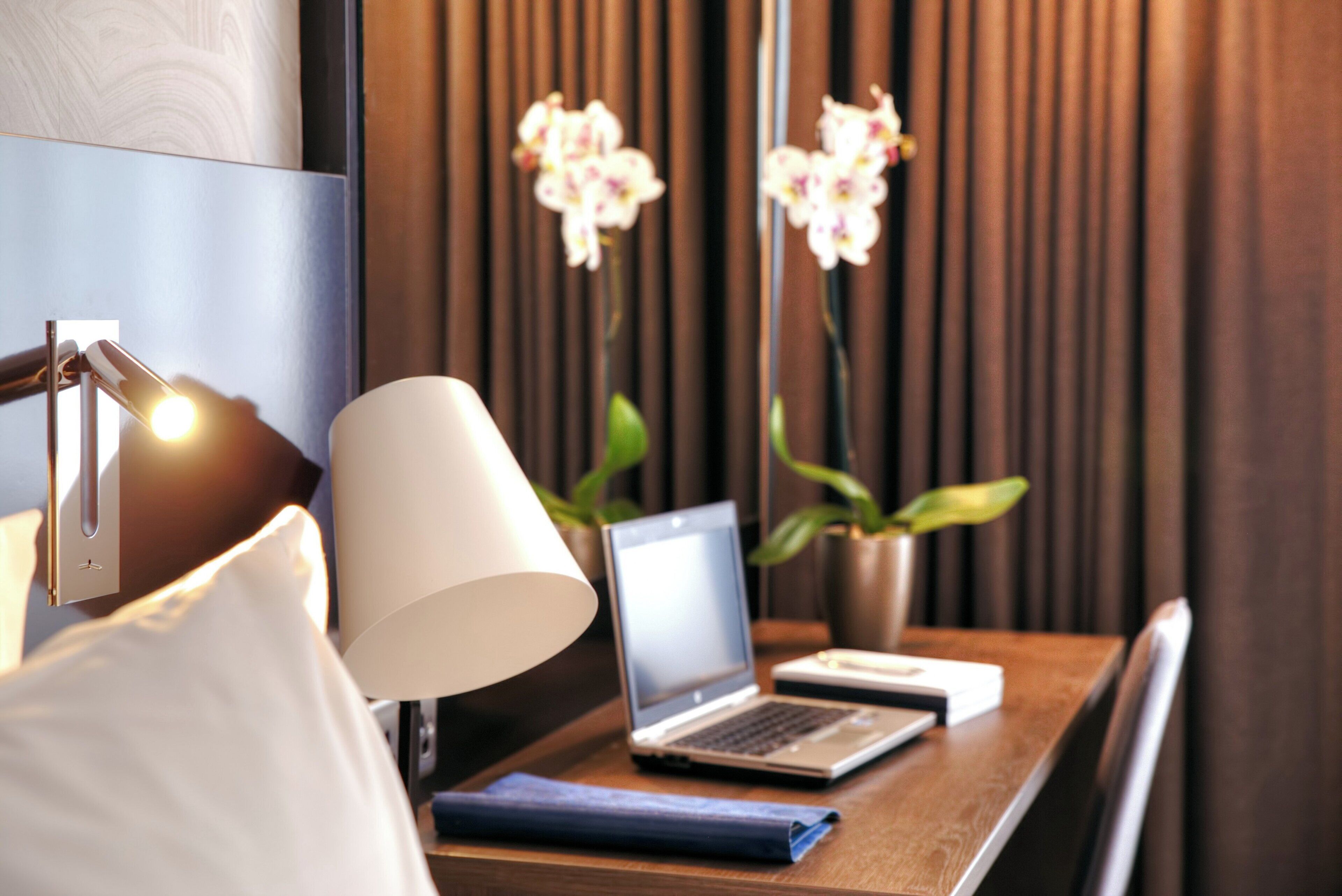 Una camera d'albergo ordinata con un portatile su un tavolo di legno, una lampada e un vaso con orchidee.