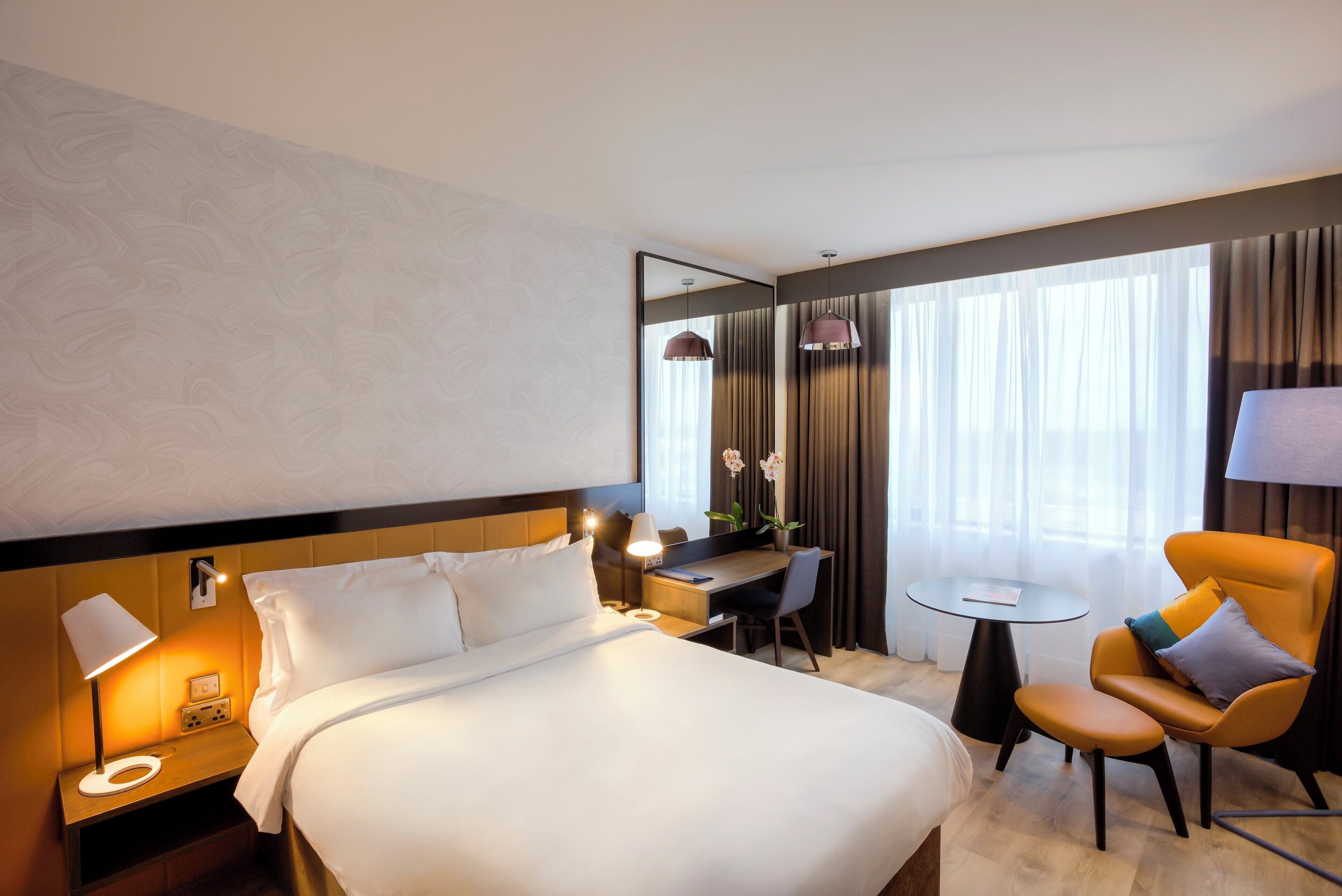 Una habitación de hotel con cama, mesita de noche, lámpara, silla, escritorio y una pequeña mesa redonda.