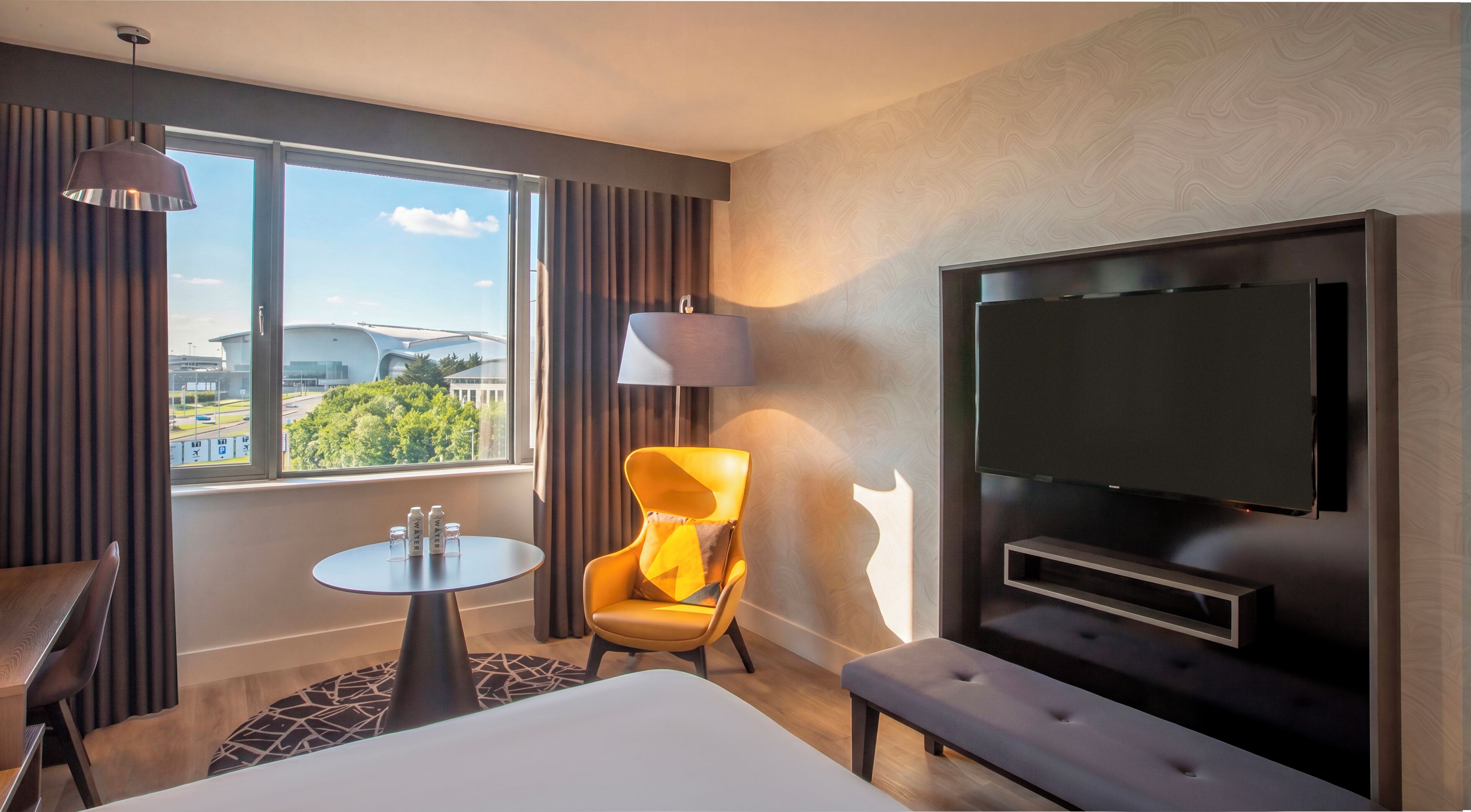 Une chambre d’hôtel avec un lit, une télévision, une chaise, une table et une fenêtre donnant sur un immeuble et des arbres.