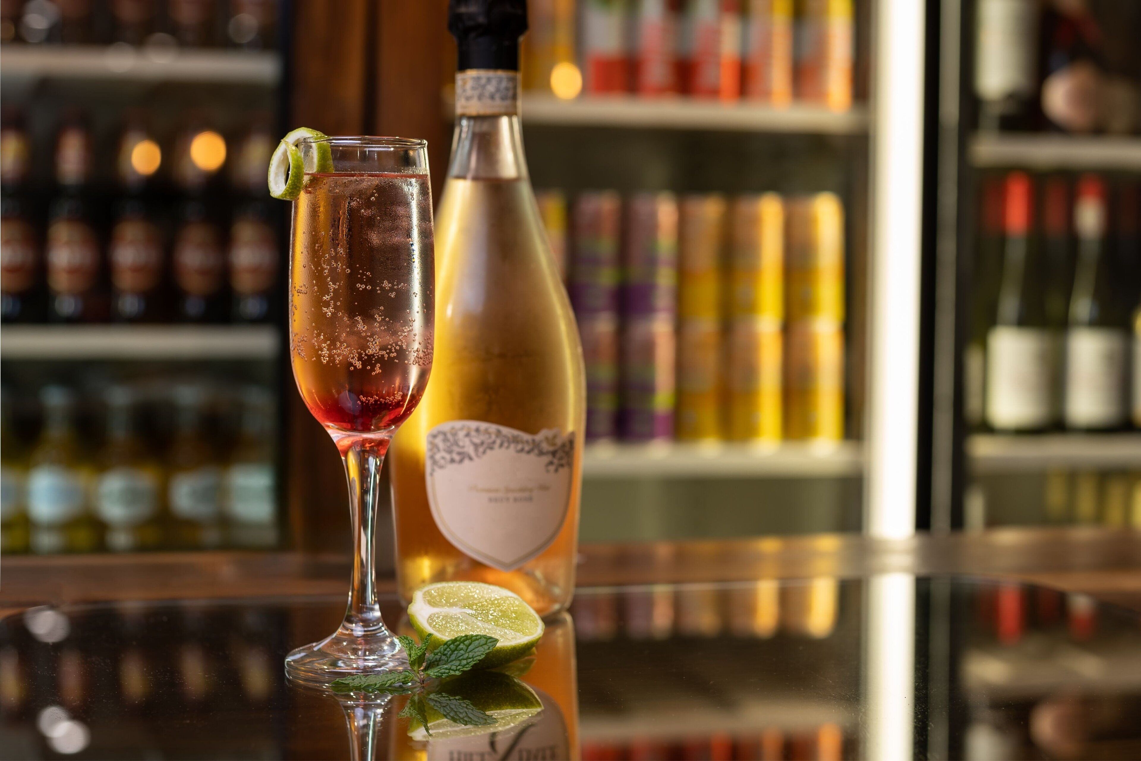 Eine Bar mit einem Glas rotem Getränk, einer Flasche Weißwein und einem Limettenschnitzchen auf einer spiegelnden Oberfläche.