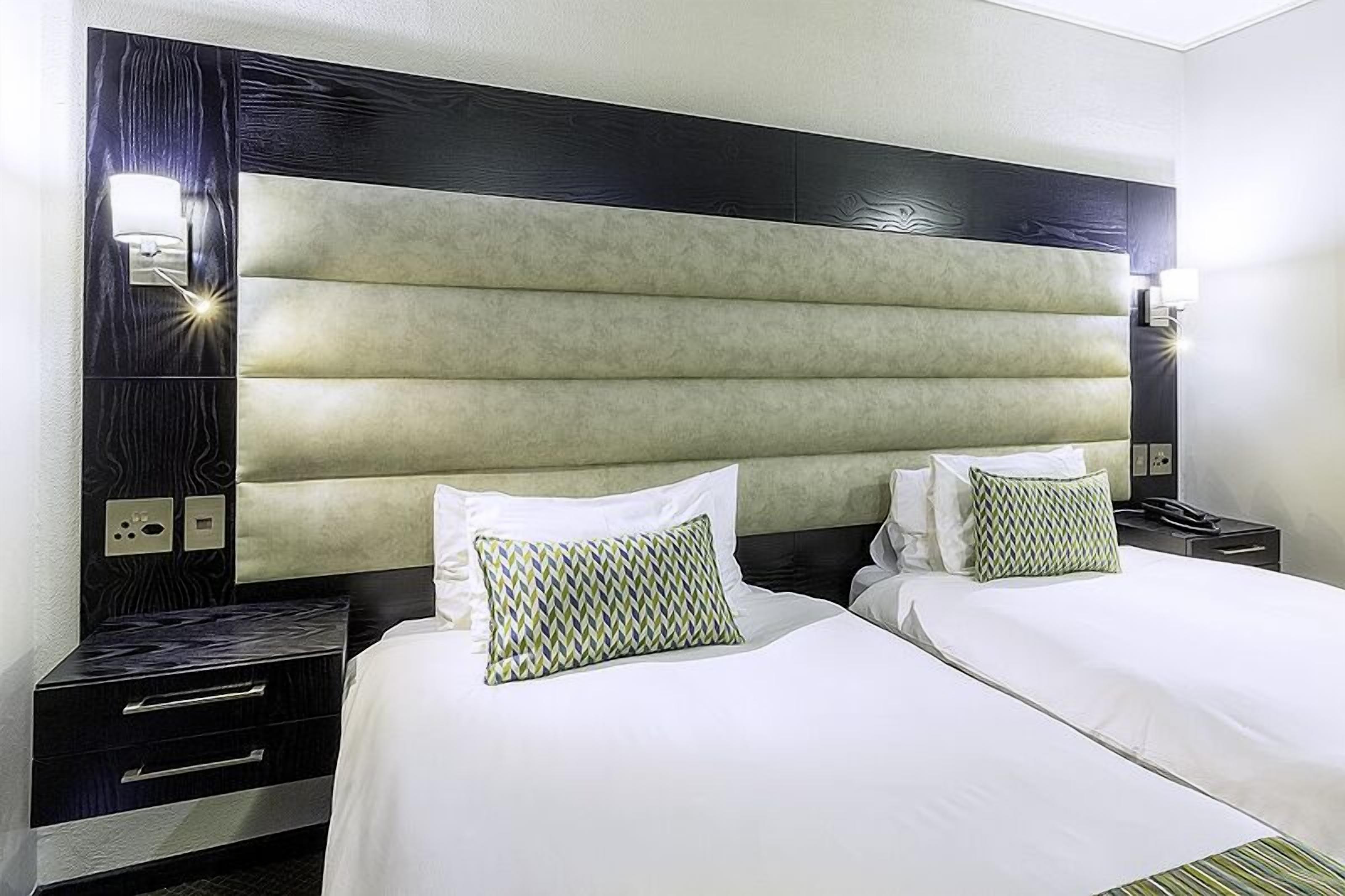 A hotel room with two beds, a headboard with a light panel, a nightstand with a drawer, and a wall socket.