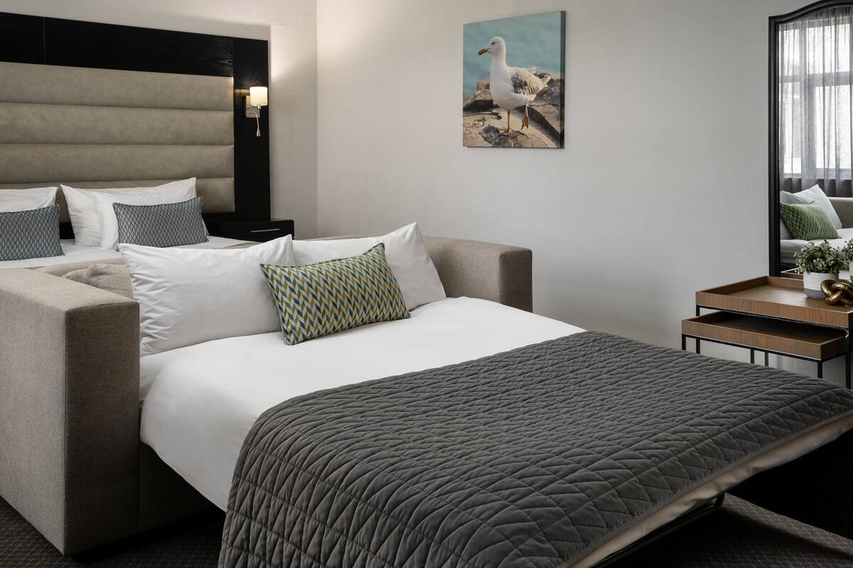 A modern hotel room with a large bed, a sofa, a bedside table with a lamp, and a framed seagull artwork on the wall.