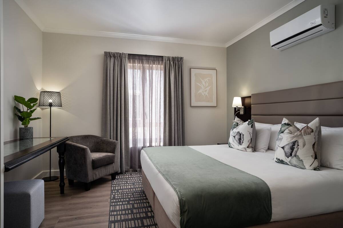 A hotel room with a bed, a desk, a chair, a lamp, a plant, and an air conditioner.