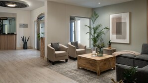 Un área de recepción moderna con mostrador, dos sillones color beige, una mesa de centro de madera y un sofá gris.