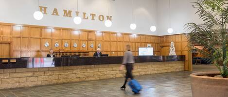 A hotel reception area with a wooden reception desk, a large sign reading "HAMILTON", and a clock above the desk.