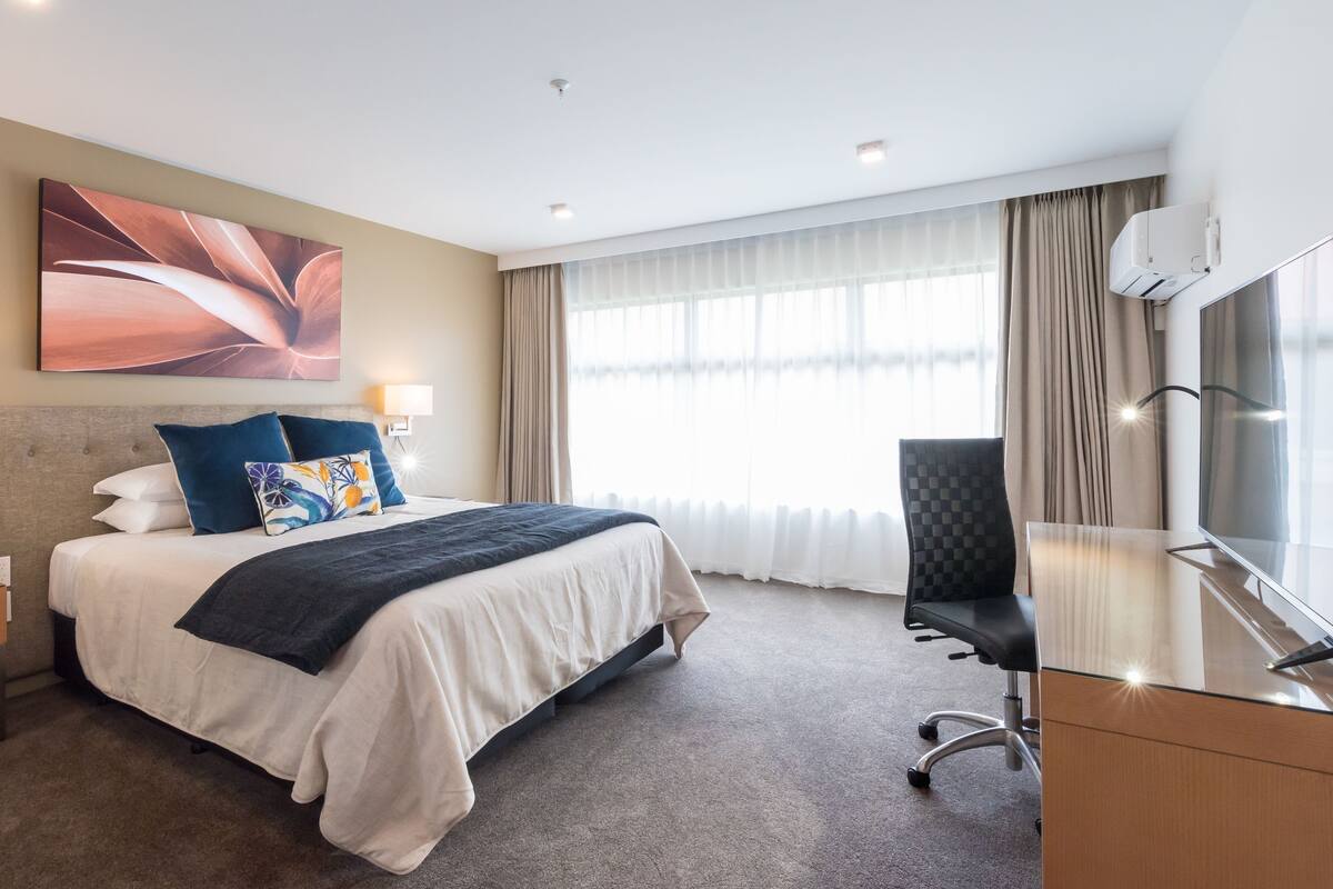 Une chambre d’hôtel avec un grand lit, un bureau avec une chaise, une télévision et une fenêtre avec des rideaux.