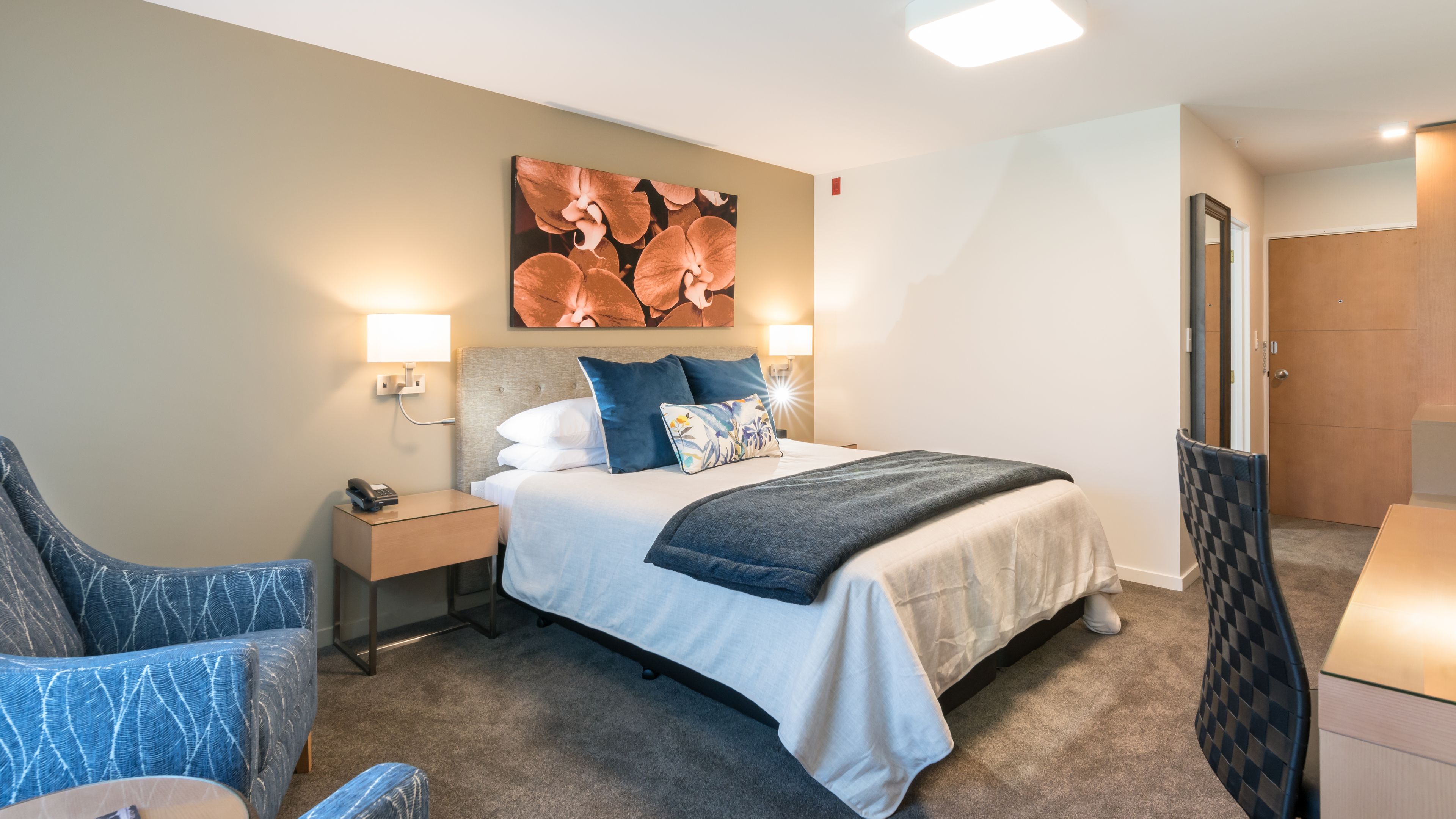 A hotel room with a bed, bedside tables, a chair, a desk, and a large floral artwork on the wall.