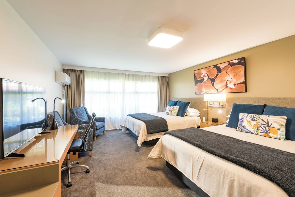 Une chambre d’hôtel avec deux lits, un bureau, une chaise, une tél évision et une grande fenêtre avec des rideaux.