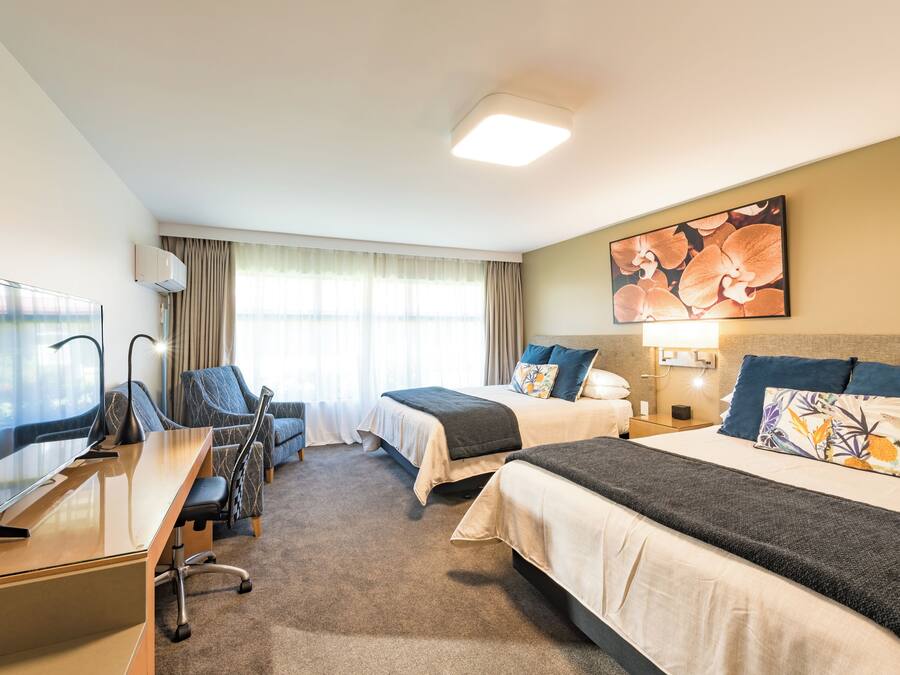 A hotel room with two beds, a desk, a chair, a TV, and a large window with curtains.