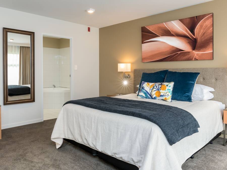 A hotel room with a bed, bedside lamps, a nightstand, a mirror, a bathroom with a sink, and a shower.