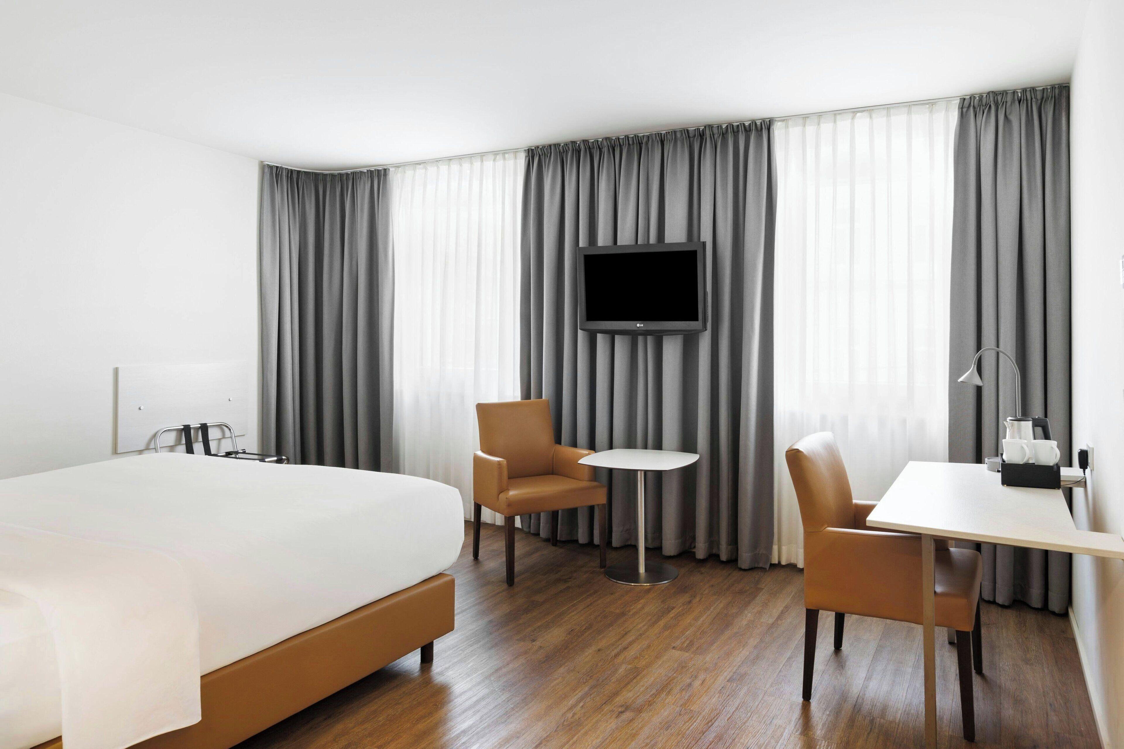 Une chambre d’hôtel avec un grand lit, deux fauteuils, une petite table, un téléviseur fixé au mur et une fenêtre avec des rideaux gris.