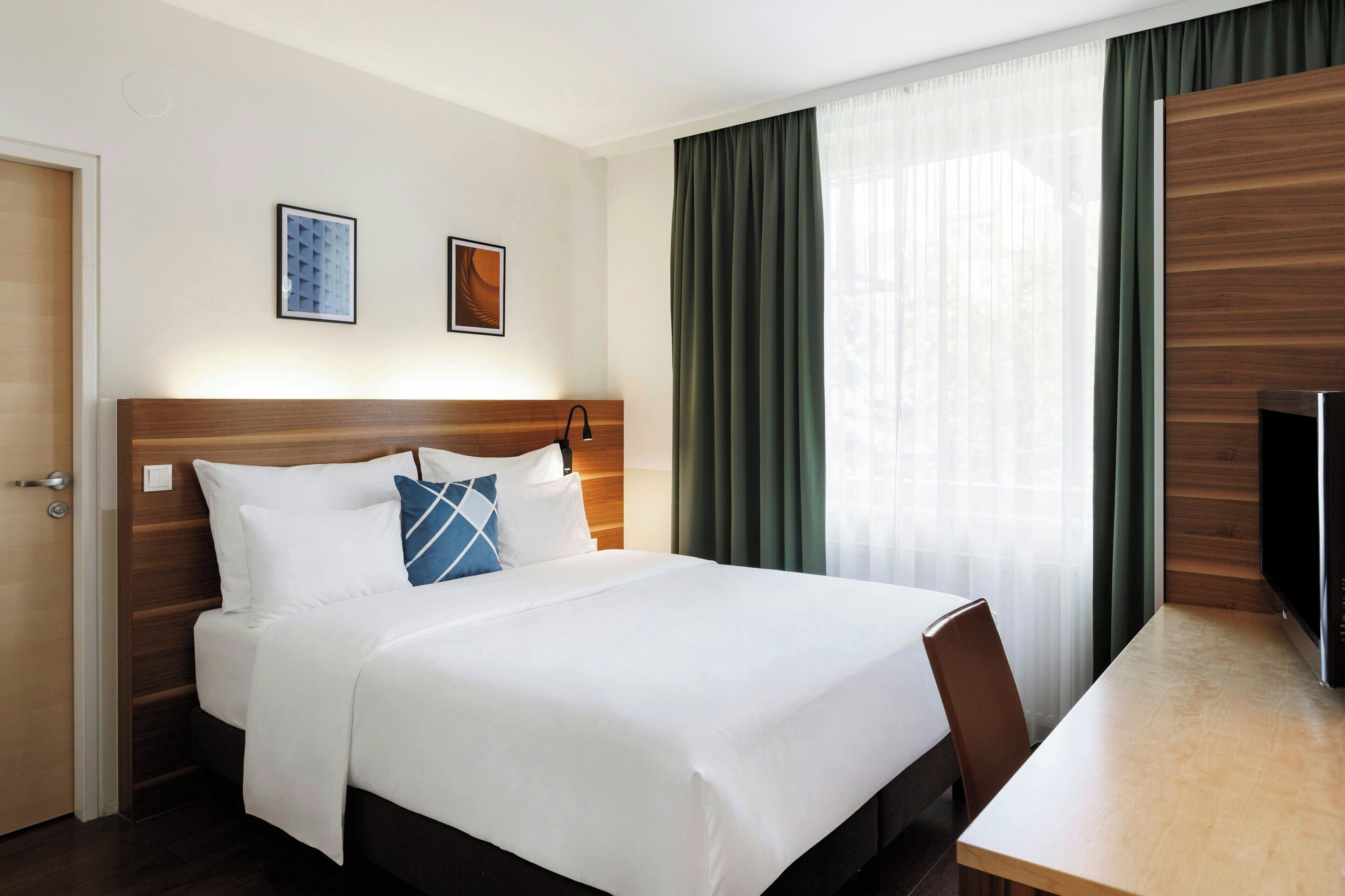 A hotel room with a wooden headboard, a bed with white linens and a blue pillow, a bedside table, and a television.