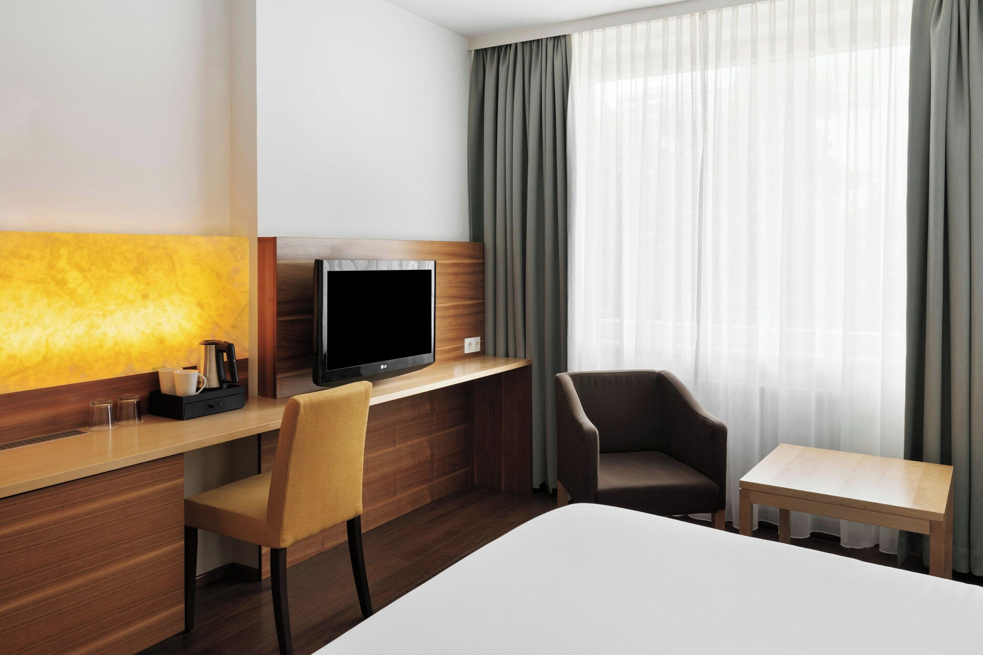 A hotel room with a bed, a desk with a flat-screen TV, a chair, a small table, and a window with curtains.