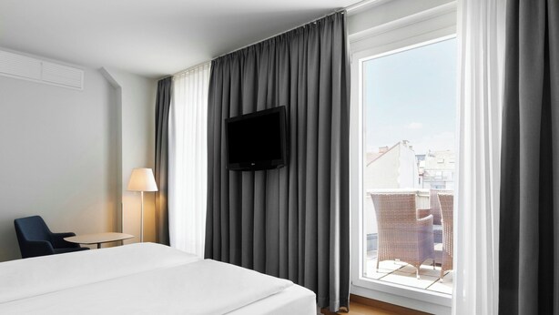 Een hotelkamer met een bed, een aan de muur bevestigde televisie, een raam met gordijnen, een stoel en een tafel.