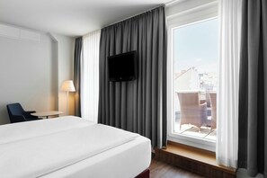 Una habitación de hotel con cama, televisor montado en la pared, ventana con cortinas, una silla y una mesa.