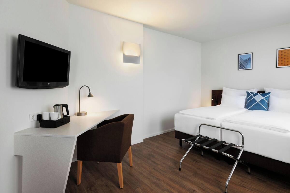 Una habitación de hotel moderna con una televisión de pantalla plana, un escritorio con una silla, una mesita de noche, una cama con cabecero y una pequeña lámpara de noche.