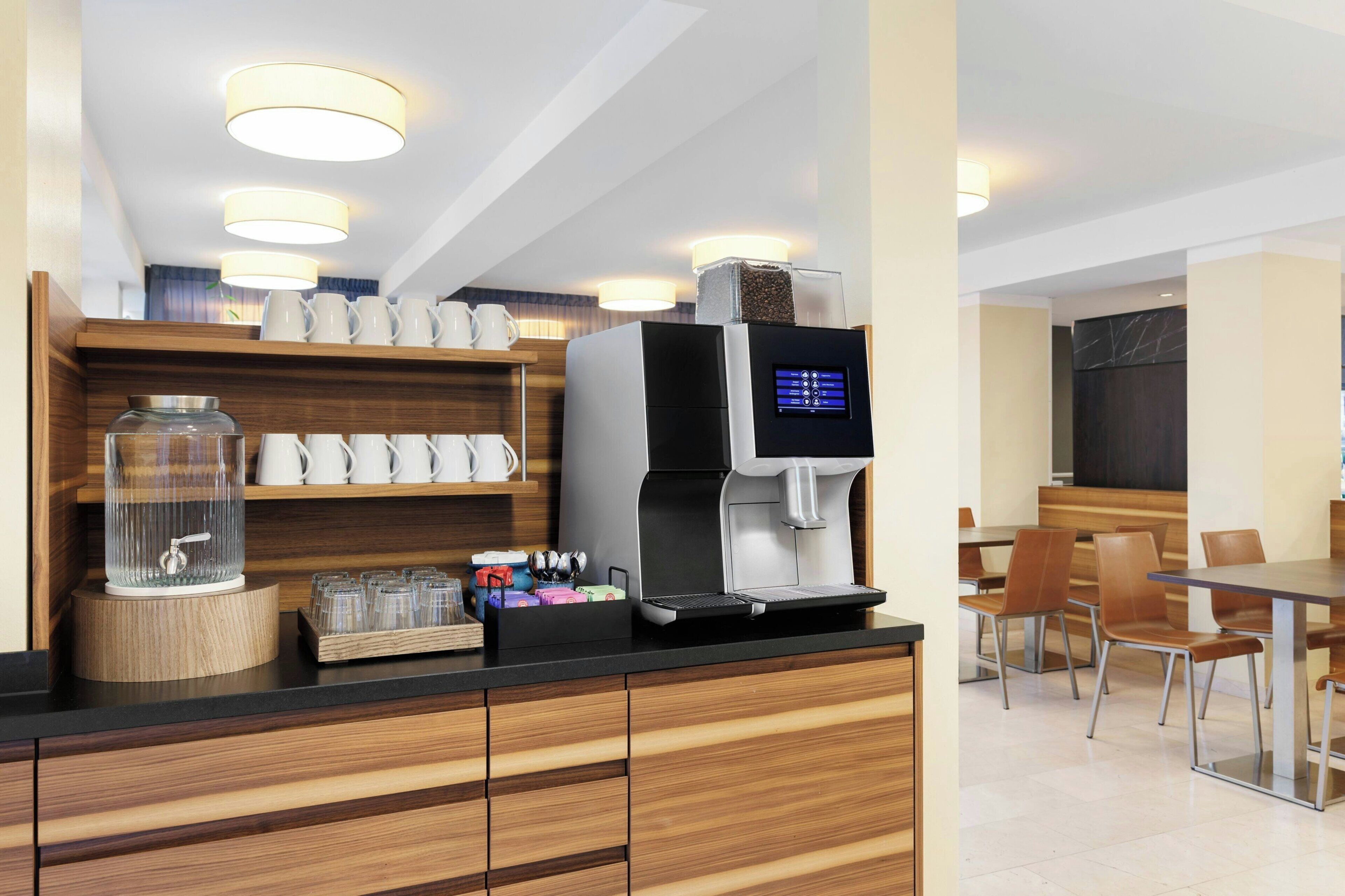 Uma estação de café moderna com uma máquina de café digital, um recipiente de vidro com torneira de metal e uma prateleira de madeira com xícaras brancas.
