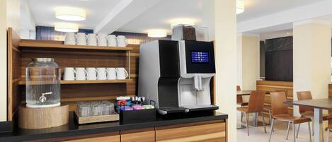 Uma estação de café moderna com uma máquina de café digital, um recipiente de vidro com torneira de metal e uma prateleira de madeira com xícaras brancas.