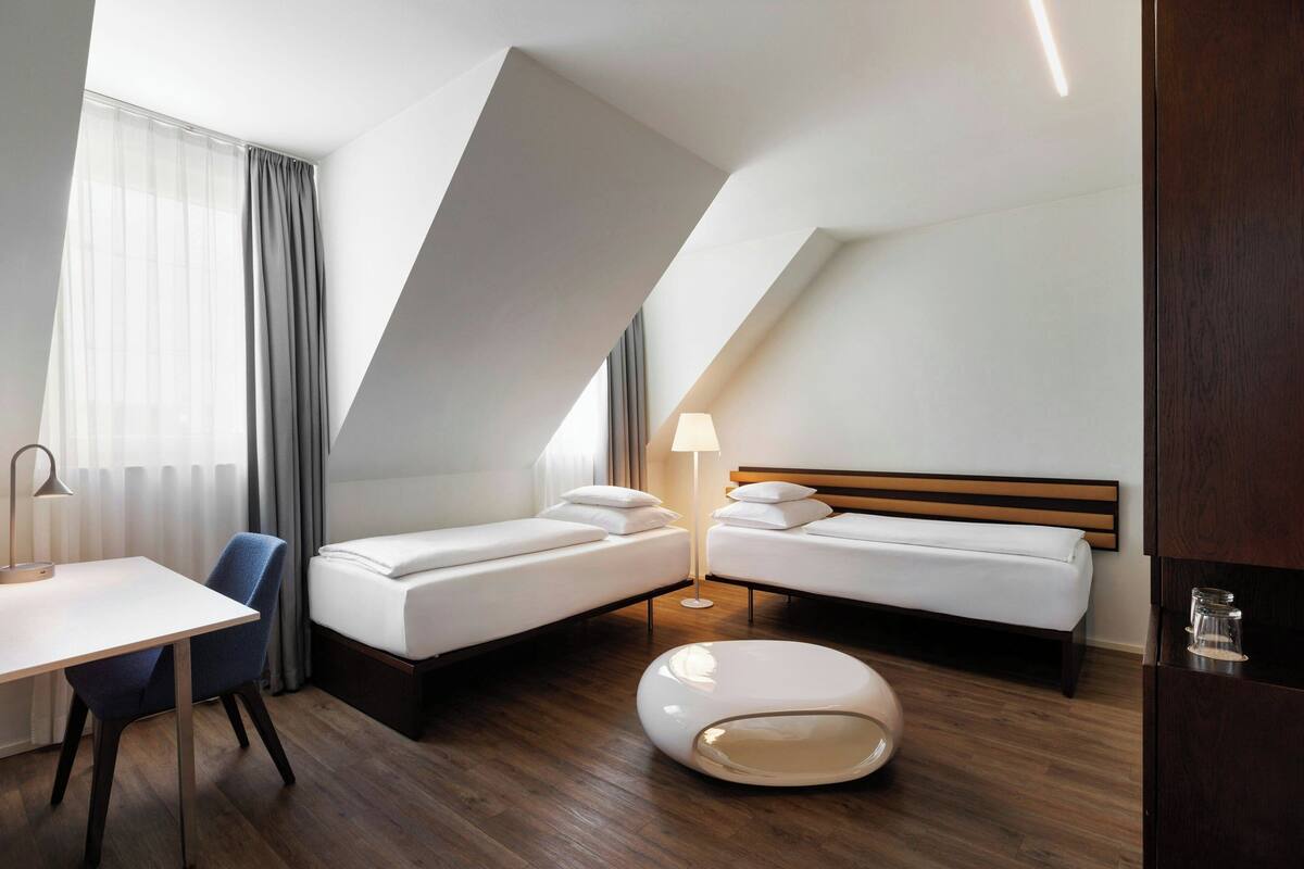 Una habitación de hotel moderna con dos camas individuales, un escritorio, una silla, un pouf redondo y una mesita de noche con lámpara.