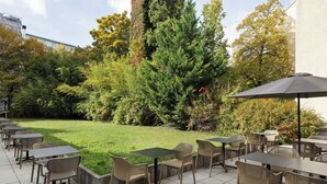 Een terras buiten met tafels en stoelen, omringd door groen en bomen.