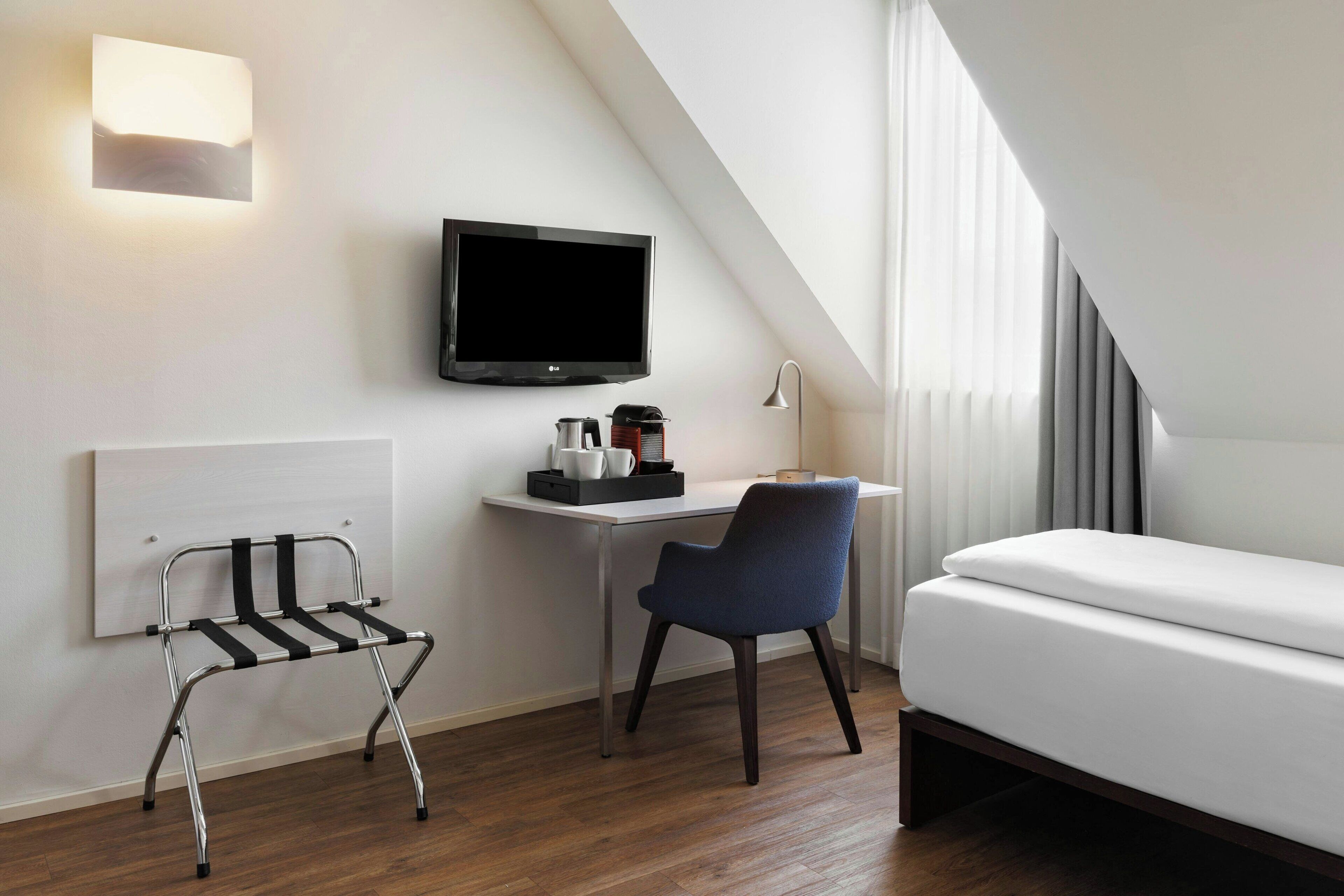 Quarto de hotel moderno com cama, escrivaninha, cadeira, televisão e mesa de cabeceira com abajur.