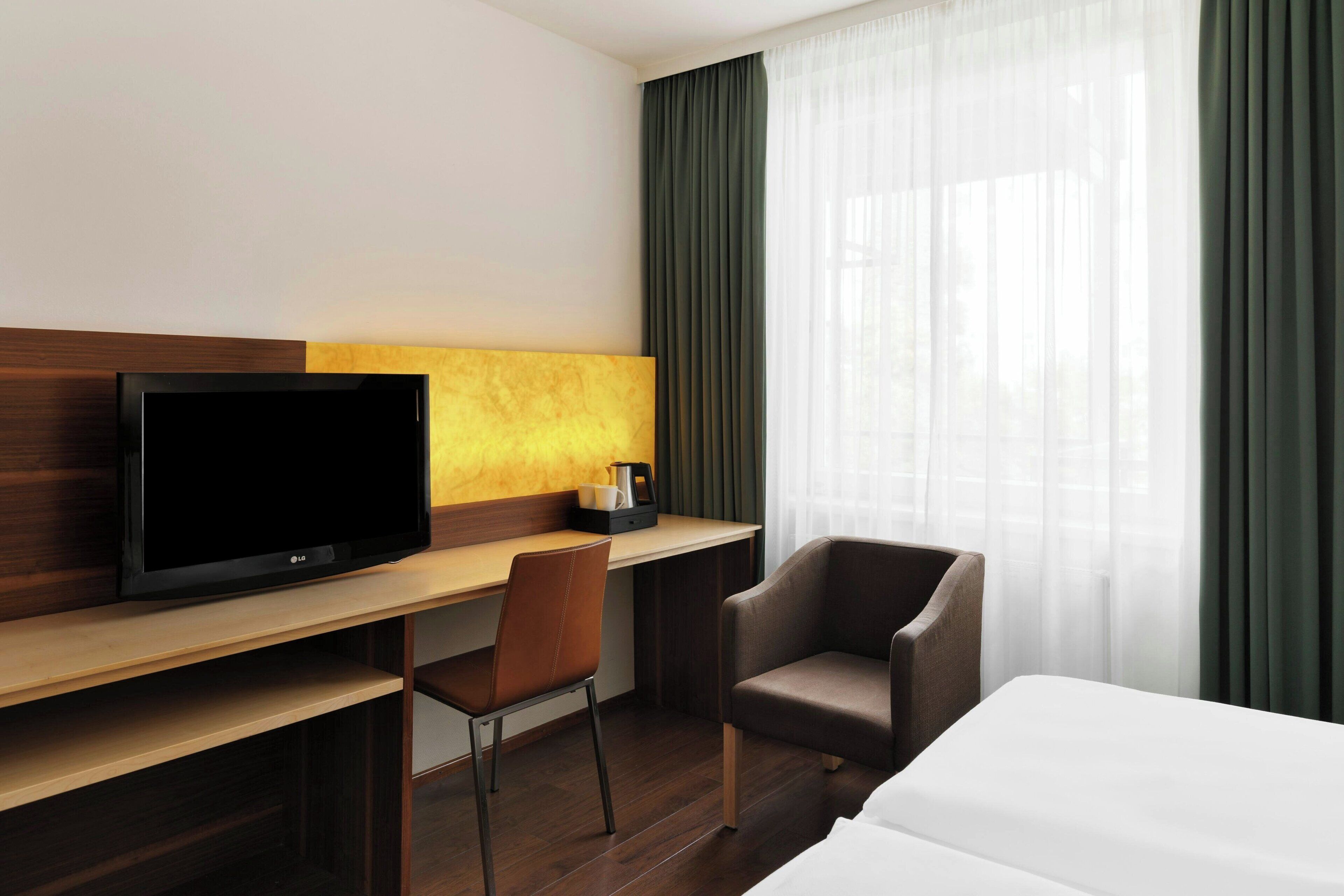 A hotel room with a wooden desk, a television, a chair, a bed, and a window with curtains.