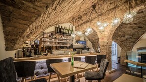 A rustic interior with exposed brick walls, a bar area with shelves of bottles, and a seating area with cushioned chairs and a wooden table.