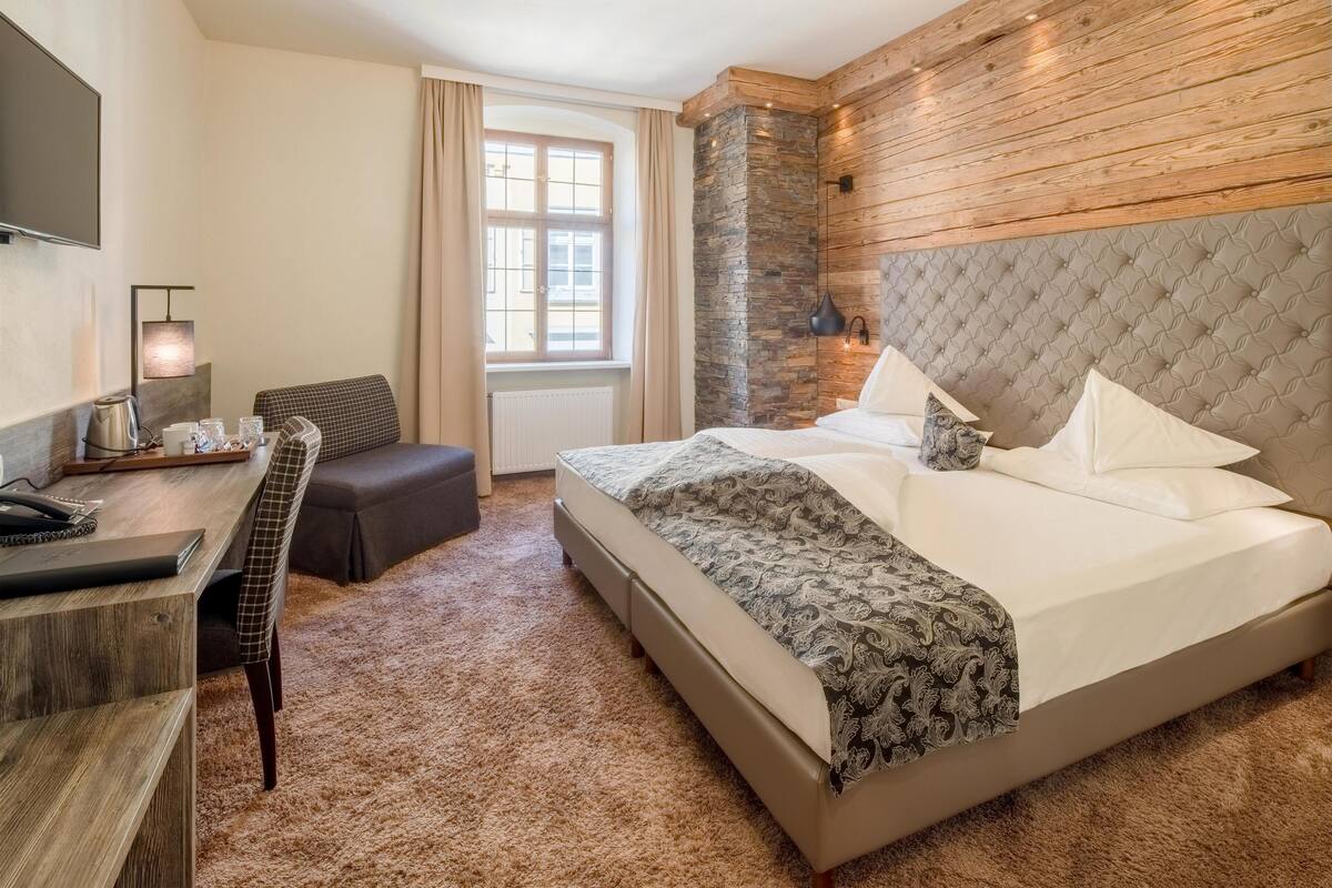A hotel room with a large bed, a desk with a telephone, a chair, a window with curtains, and a stone accent wall.