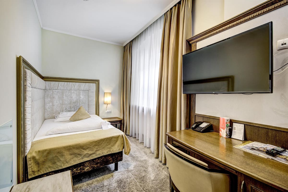 A hotel room with a bed, a desk with a telephone, a flat-screen TV, and a window with curtains.