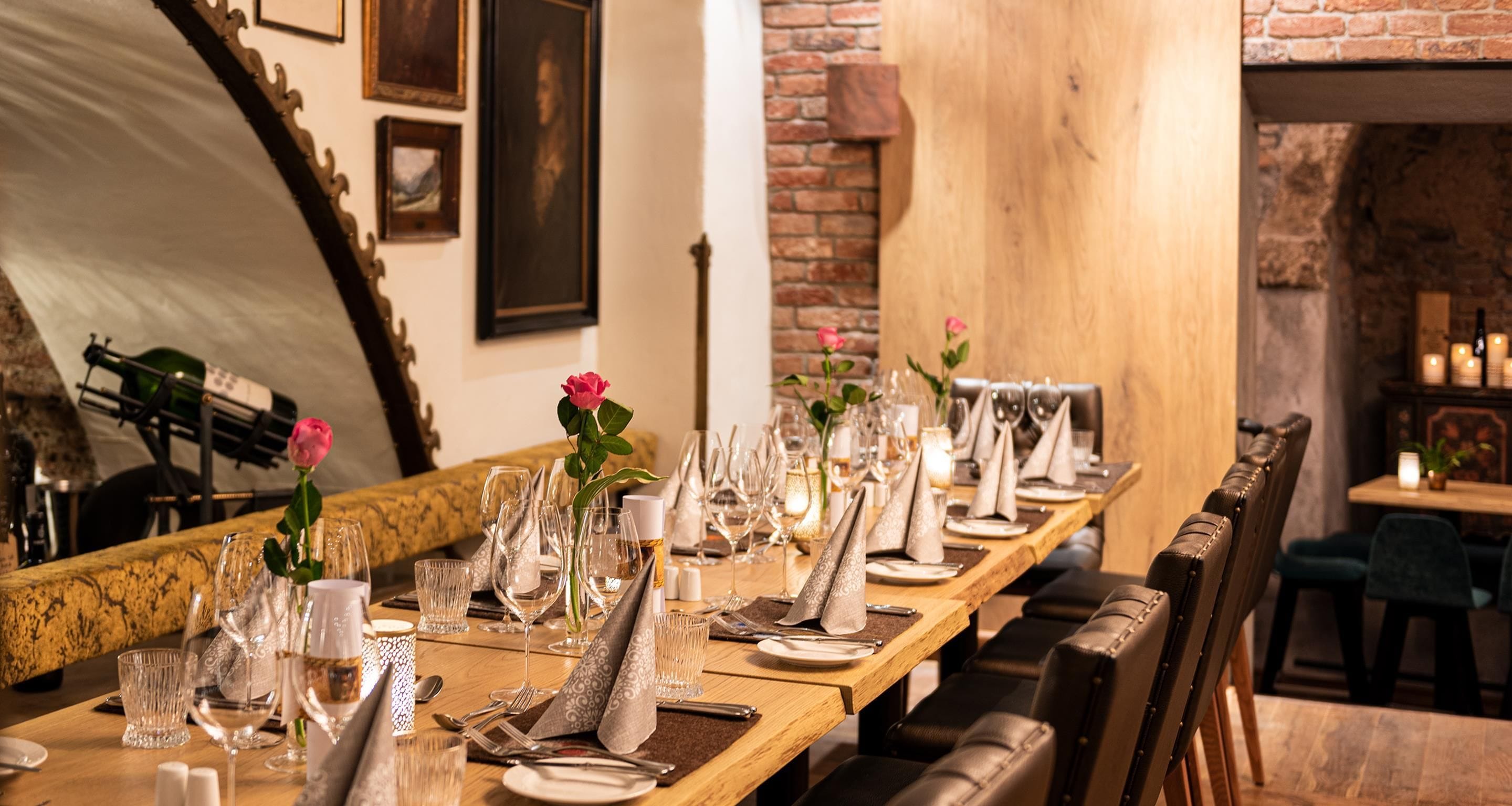 A long dining table set for a meal with multiple chairs, a brick wall, and a wooden staircase in the background.