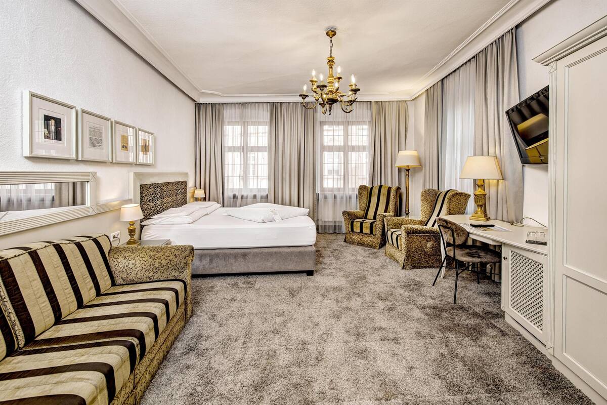 A hotel room with a bed, a sofa, a desk, a chair, a chandelier, and curtains.