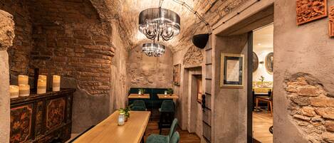 A narrow, rustic dining area with a wooden table and green upholstered chairs, a chandelier, and candlelit ambiance.