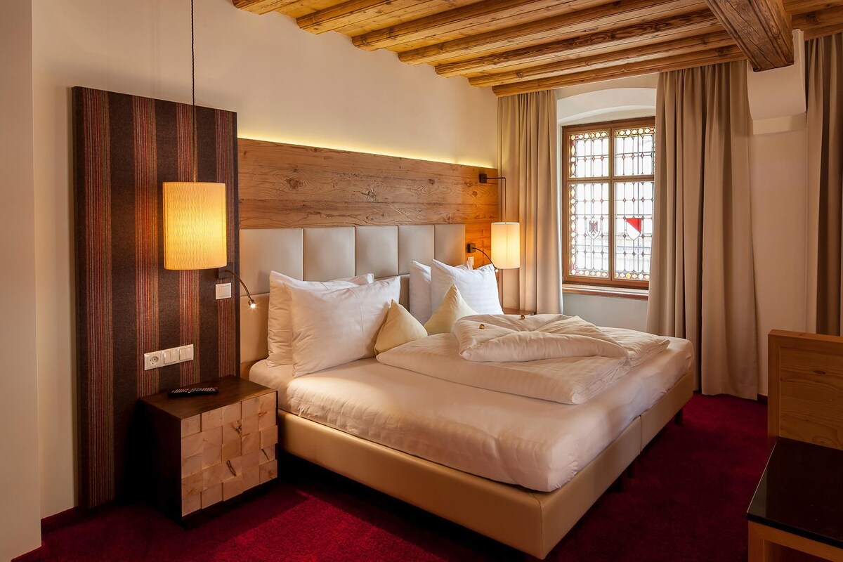 Une chambre d’hôtel avec un grand lit, une tête de lit en bois, une table de chevet avec une lampe, une commode en bois et une fenêtre avec des rideaux.