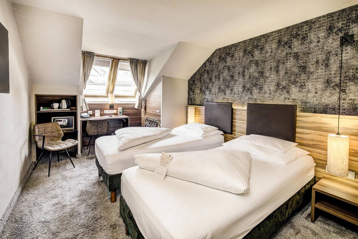 A hotel room with two beds, a desk, a chair, a shelf with items, and a window with curtains.