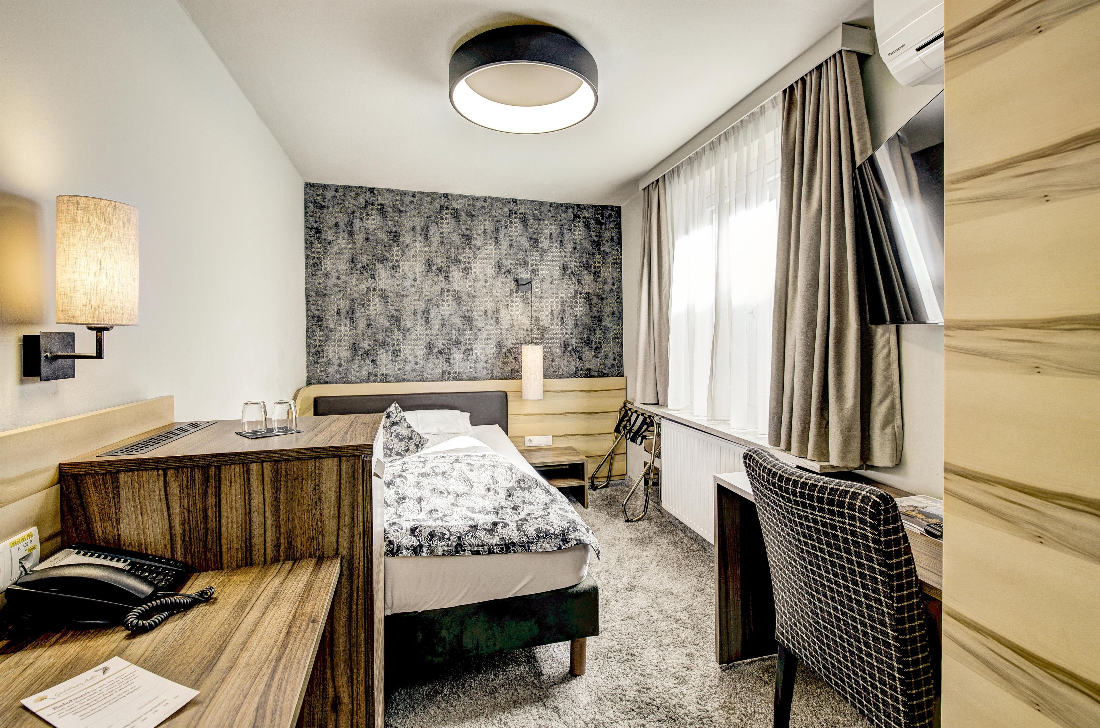 A hotel room with a bed, bedside table, lamp, and a view of the outside.
