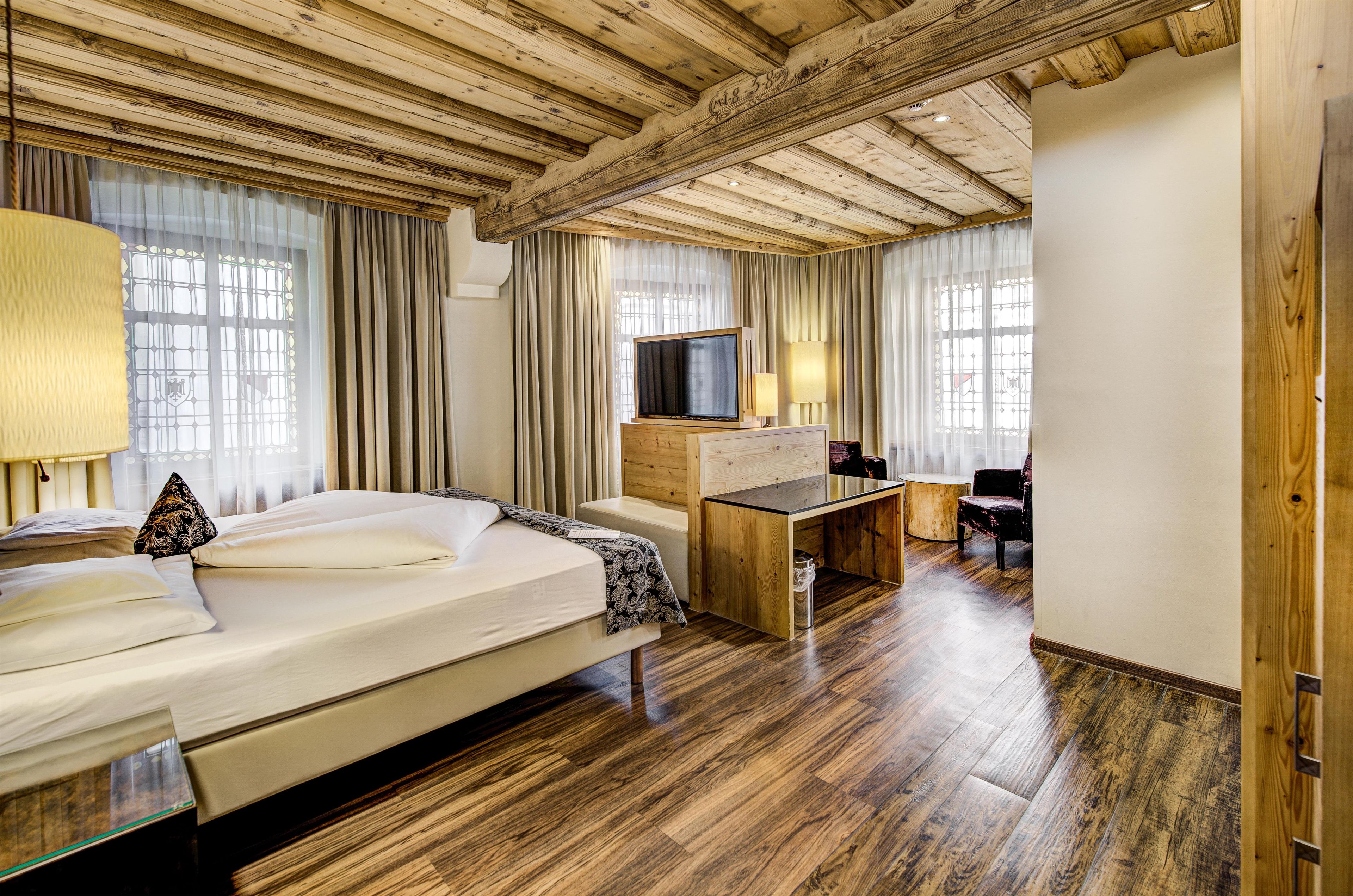 A hotel room with a wooden ceiling, a bed, a desk, a television, and a window with curtains.