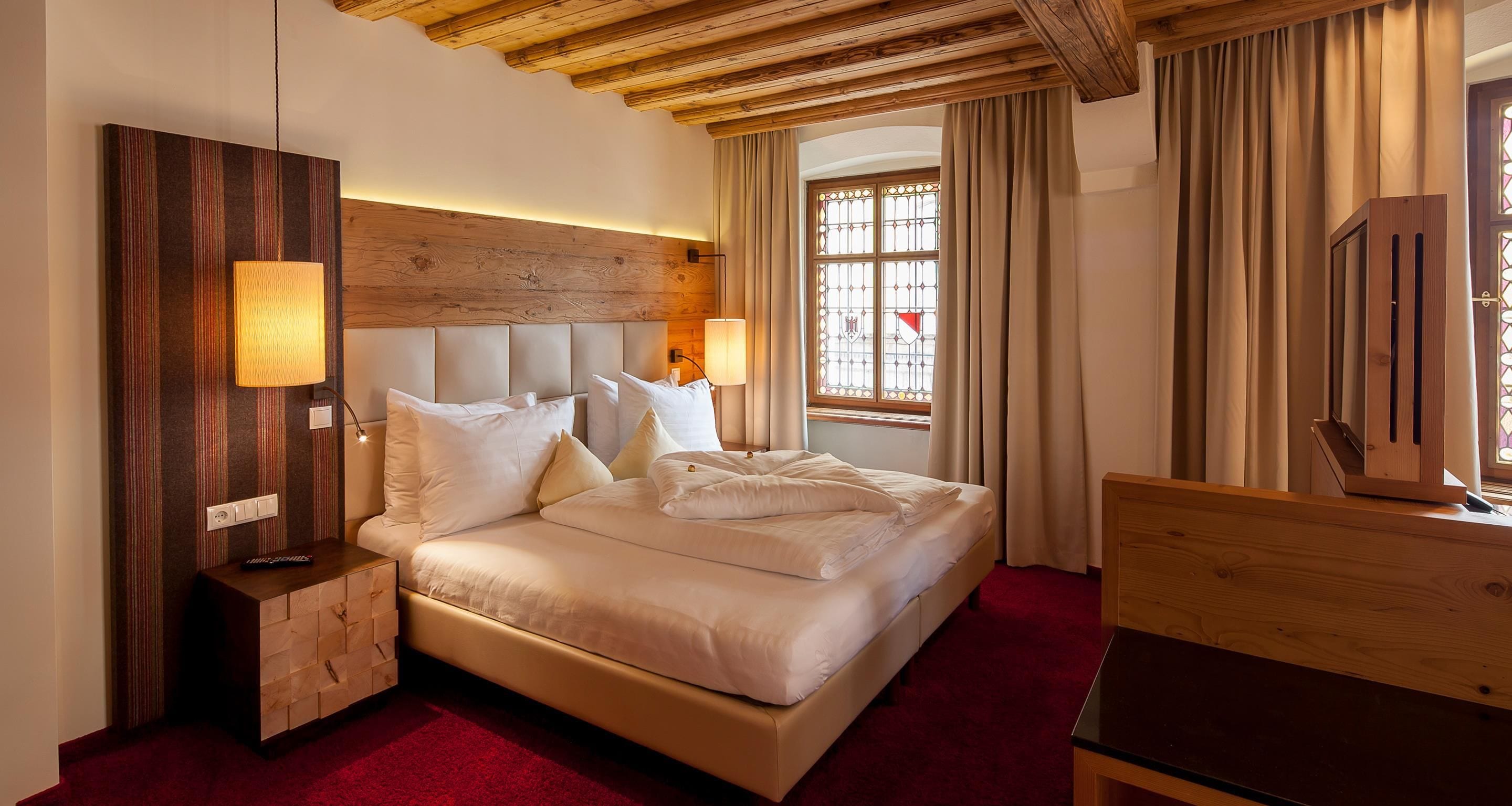 Habitación de hotel con una cama grande, cabecero de madera, una mesita de noche con lámpara, cómoda de madera y una ventana con cortinas.