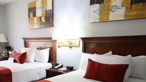 A hotel room with two beds, each with white linens and red accents, a wooden headboard, a nightstand with a lamp, and artwork on the walls.