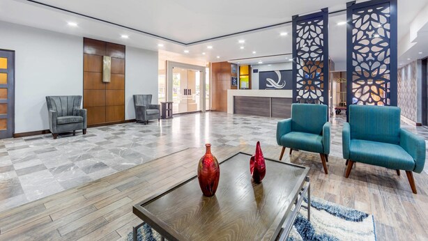 A spacious lobby with a checkered floor, two blue armchairs, a wooden coffee table with two red vases, and a decorative wall panel.