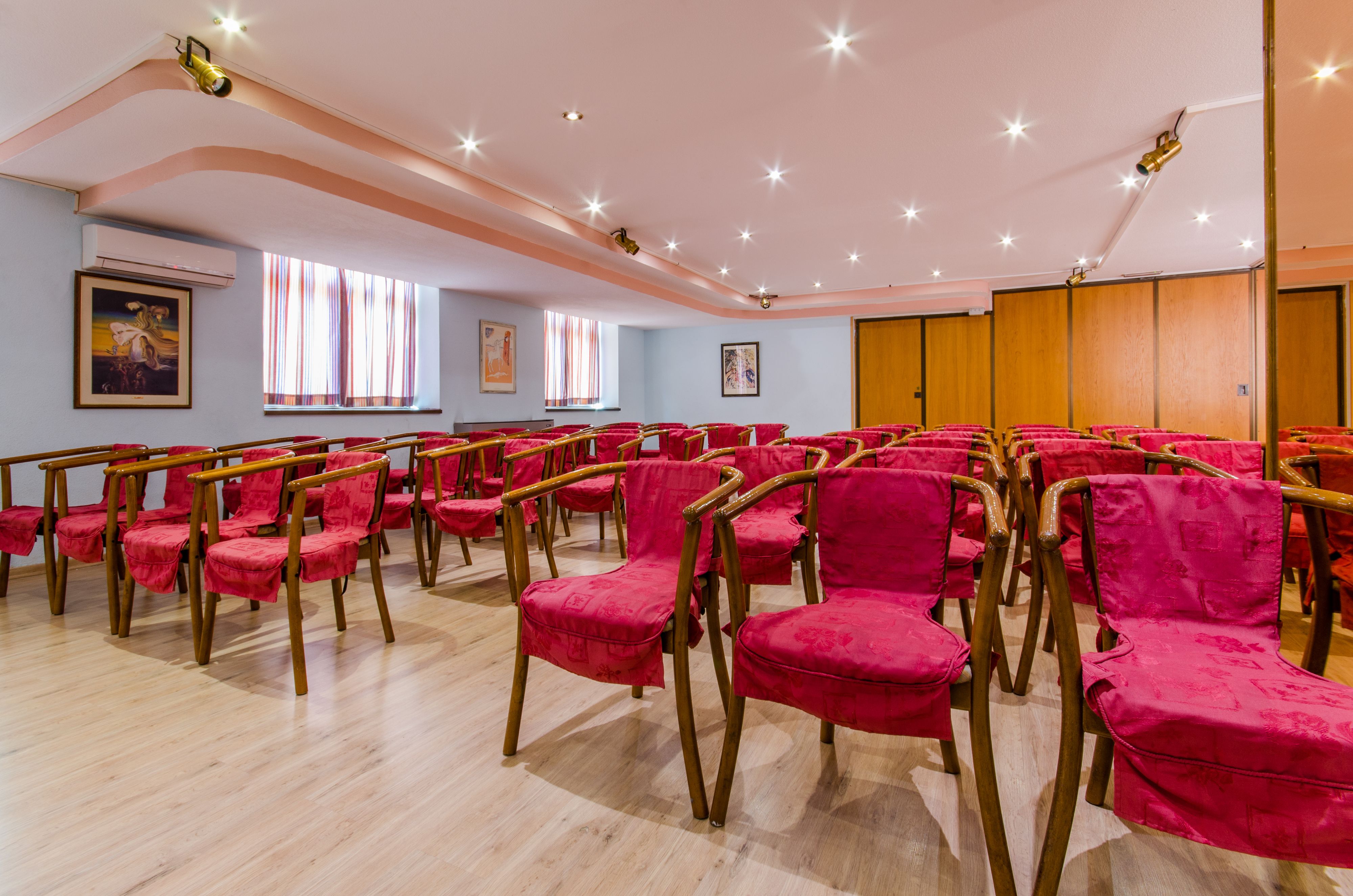 Una sala conferenze con file di sedie rosse e tavoli di legno, ampie finestre con tende a righe e pannelli murali in legno.