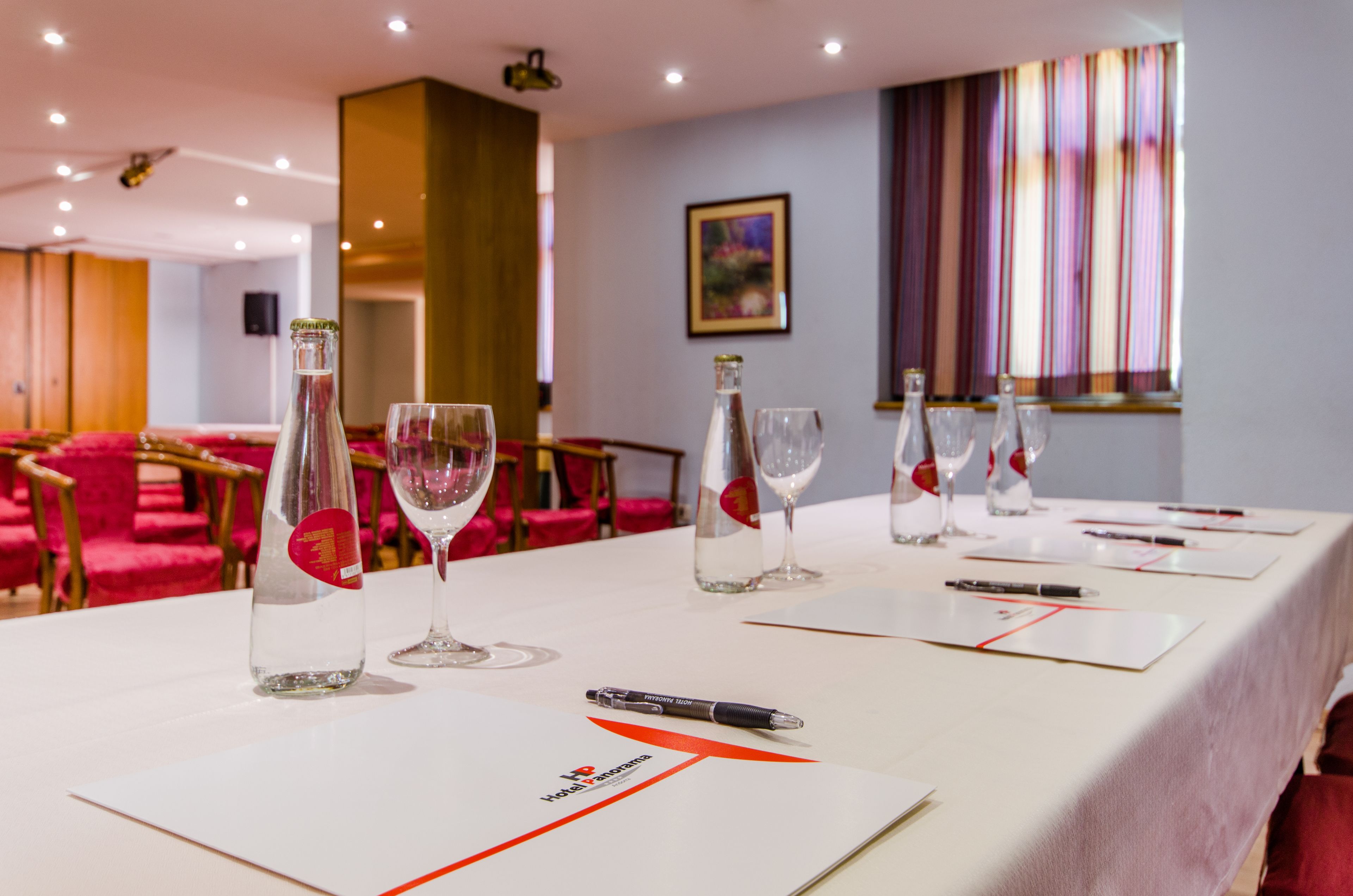 A conference table set with water bottles, glasses, and notepads in a room with red chairs and a painting on the wall.